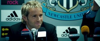 Movie still from “Goal! The Dream Begins” (2005), directed by Michael Winterbottom – A person sitting at a microphone in a room; Close Up shot, Over the shoulder angle