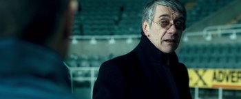 Movie still from “Goal! The Dream Begins” (2005), directed by Michael Winterbottom – An older man wearing a black coat and a black tie; Close Up shot, Over the shoulder angle