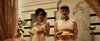 Movie still from “Gods of Egypt” (2016), directed by Alex Proyas – Two young women dressed in ancient egyptian clothing holding plates of food in front of an egyptian wall; Medium shot, Over the shoulder angle