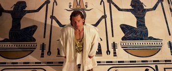 Movie still from “Gods of Egypt” (2016), directed by Alex Proyas – A man wearing a robe standing in front of an egyptian mural; Medium shot, Overhead angle