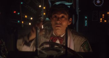 Movie still from “Godzilla vs. Mechagodzilla II” (1993), directed by Kazuki Ômori – A man in a hat and a bow tie is sitting in front of a steering wheel; Close Up shot, Overhead angle