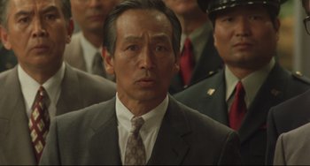 Movie still from “Godzilla vs. Mechagodzilla II” (1993), directed by Kazuki Ômori – A man in a suit and tie standing in front of a group of men; Close Up shot, Low angle