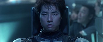 Movie still from “Godzilla: Final Wars” (2004), directed by Koji Hashimoto – A man in a black jacket is sitting down; Close Up shot, Low angle