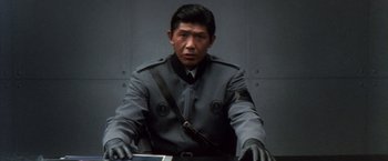 Movie still from “Godzilla: Final Wars” (2004), directed by Koji Hashimoto – A man in a uniform sitting at a table; Medium shot, Low angle