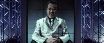 Movie still from “Godzilla: Final Wars” (2004), directed by Koji Hashimoto – A man wearing a suit and tie sitting in front of a television screen; Close Up shot, Low angle