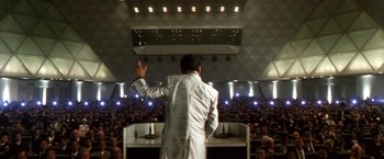 Movie still from “Godzilla: Final Wars” (2004), directed by Koji Hashimoto – A man in a white suit is giving a speech to an audience; Extreme Wide shot, High angle