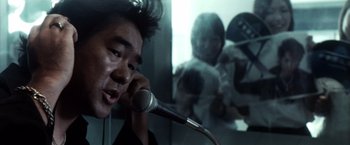Movie still from “Godzilla: Final Wars” (2004), directed by Koji Hashimoto – A man talking into a microphone in front of a mirror; Close Up shot, Over the shoulder angle