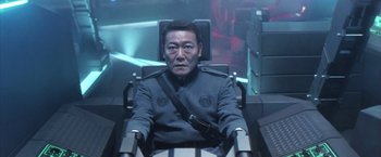 Movie still from “Godzilla: Final Wars” (2004), directed by Koji Hashimoto – A man sitting in a chair in front of a screen; Medium shot, High angle