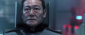 Movie still from “Godzilla: Final Wars” (2004), directed by Koji Hashimoto – An older man wearing a black shirt and a gray jacket; Close Up shot, Low angle