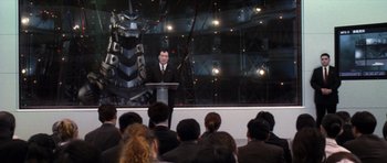 Movie still from “Godzilla: Tokyo S.O.S.” (2003), directed by Takao Okawara – A man in a black suit is speaking at a podium; Extreme Wide shot, High angle