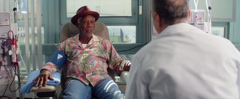 Movie still from “Going in Style” (2017), directed by Zach Braff – An older man sitting in a chair talking to a doctor; Medium shot, Over the shoulder angle