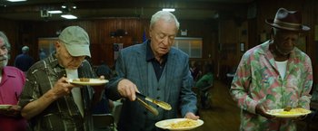 Movie still from “Going in Style” (2017), directed by Zach Braff – An older man holding a knife and a plate of food; Medium shot, Low angle