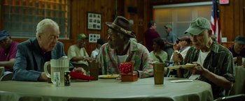 Movie still from “Going in Style” (2017), directed by Zach Braff – An older man sitting at a table with a hat on; Medium shot, Over the shoulder angle