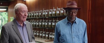 Movie still from “Going in Style” (2017), directed by Zach Braff – A man wearing a hat standing in front of a wall filled with jars; Medium shot, Over the shoulder angle