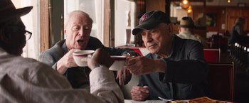Movie still from “Going in Style” (2017), directed by Zach Braff – Two older men sitting at a table and one of them is holding a plate; Medium shot, Low angle