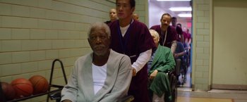 Movie still from “Going in Style” (2017), directed by Zach Braff – An older man sitting in a wheelchair in front of a group of people; Medium shot, High angle