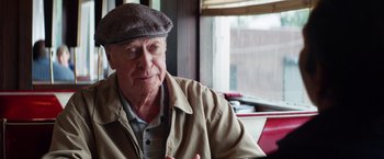 Movie still from “Going in Style” (2017), directed by Zach Braff – An older man wearing a hat sitting at a table in front of a window; Close Up shot, Over the shoulder angle