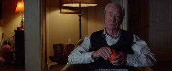 Movie still from “Going in Style” (2017), directed by Zach Braff – An older man holding an orange sitting in a chair; Medium shot, Over the shoulder angle