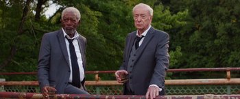 Movie still from “Going in Style” (2017), directed by Zach Braff – A man in a suit and tie standing next to another man in a suit and tie; Medium shot, Low angle