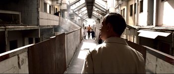 Movie still from “Gomorrah” (2008), directed by Matteo Garrone – A man standing on a walkway with people walking around; Wide shot, Over the shoulder angle