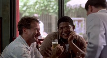 Movie still from “Good Morning, Vietnam” (1987), directed by Barry Levinson – Two men are sitting at a table and one of them is holding a drink; Medium shot, Low angle
