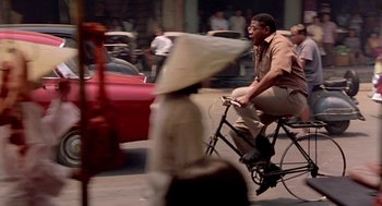 Movie still from “Good Morning, Vietnam” (1987), directed by Barry Levinson – A man riding a bike down a street past a fire hydrant; Wide shot, Over the shoulder angle