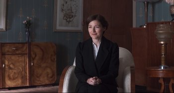 Movie still from “Goodbye Christopher Robin” (2017), directed by Simon Curtis – A woman in a suit sitting in a chair in a room; Medium shot, Over the shoulder angle