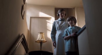Movie still from “Goodbye Christopher Robin” (2017), directed by Simon Curtis – A woman and a young boy standing in a room next to a lamp; Medium shot, Low angle