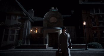 Movie still from “Goodbye Christopher Robin” (2017), directed by Simon Curtis – A woman in a hat holding a briefcase in front of a building at night; Wide shot, Low angle