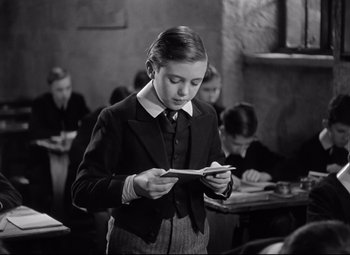 Movie still from “Goodbye, Mr. Chips” (1939), directed by Sam Wood – A young boy reading a book in front of a group of people; Medium shot, Low angle