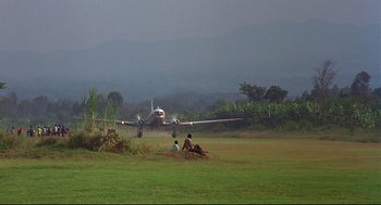 Movie still from “Gorillas in the Mist” (1988), directed by Michael Apted – An airplane taking off from a runway with two people sitting on the ground; Extreme Wide shot, High angle