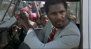 Movie still from “Gorillas in the Mist” (1988), directed by Michael Apted – A man in a suit and tie in front of a group of people; Close Up shot, Over the shoulder angle