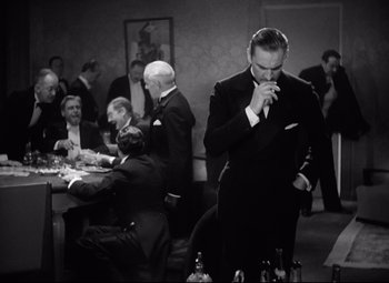 Movie still from “Grand Hotel” (1932), directed by Edmund Goulding – Black and white photograph of a man smoking a cigarette in front of a group of people; Medium shot, High angle