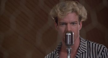 Movie still from “Great Balls of Fire!” (1989), directed by Jim McBride – A man with blonde curly hair is holding a microphone up to his mouth; Close Up shot, Low angle