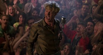 Movie still from “Great Balls of Fire!” (1989), directed by Jim McBride – A man singing into a microphone in front of a crowd; Medium shot, Low angle