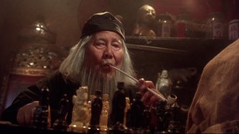 Movie still from “Gremlins 2: The New Batch” (1990), directed by Joe Dante – An old man smoking a pipe in front of a group of figurines; Close Up shot, High angle
