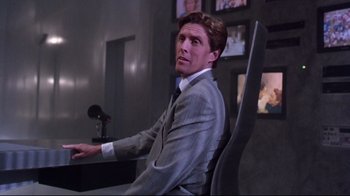 Movie still from “Gremlins 2: The New Batch” (1990), directed by Joe Dante – A man in a suit sitting in a chair in front of a microphone; Medium shot, Low angle