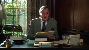 Movie still from “Gross Anatomy” (1989), directed by Thom Eberhardt – An older man sitting at a desk holding papers; Medium shot, Low angle