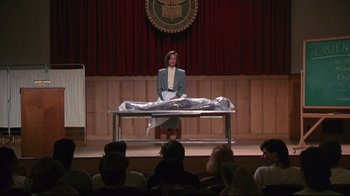 Movie still from “Gross Anatomy” (1989), directed by Thom Eberhardt – A woman sitting at a table in front of an audience; Wide shot, High angle