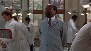 Movie still from “Gross Anatomy” (1989), directed by Thom Eberhardt – A man in a lab coat and tie standing in front of a group of people; Medium shot, Low angle