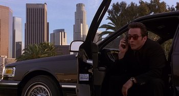 Movie still from “Grosse Pointe Blank” (1997), directed by George Armitage – A man sitting in the drivers seat of a car talking on a cell phone; Medium shot, Low angle