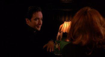 Movie still from “Grosse Pointe Blank” (1997), directed by George Armitage – A man sitting at a table in front of a lamp; Close Up shot, Over the shoulder angle