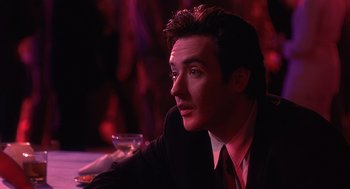 Movie still from “Grosse Pointe Blank” (1997), directed by George Armitage – A man sitting at a table with a drink in front of him; Close Up shot, Over the shoulder angle