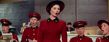 Movie still from “Guys and Dolls” (1955), directed by Joseph L. Mankiewicz – A woman in a red coat and black hat singing; Medium shot, Low angle