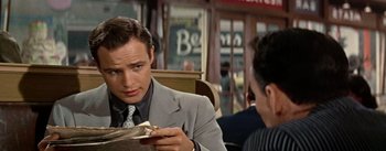 Movie still from “Guys and Dolls” (1955), directed by Joseph L. Mankiewicz – A man in a suit and tie sitting in front of a newspaper; Close Up shot, Over the shoulder angle
