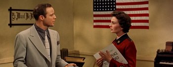 Movie still from “Guys and Dolls” (1955), directed by Joseph L. Mankiewicz – A woman holding a sign talking to a man in front of an american flag; Medium shot, Over the shoulder angle