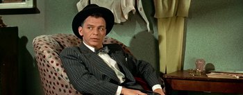 Movie still from “Guys and Dolls” (1955), directed by Joseph L. Mankiewicz – A man wearing a suit and hat sitting in a chair; Medium shot, Over the shoulder angle