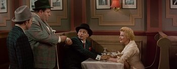 Movie still from “Guys and Dolls” (1955), directed by Joseph L. Mankiewicz – A man in a suit and a hat sitting at a table with two women; Medium shot, Over the shoulder angle