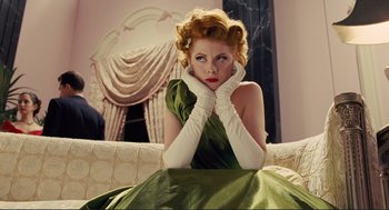 Movie still from “Hail, Caesar!” (2016), directed by Ethan Coen – A woman in a green dress and white gloves sitting on a couch; Medium shot, Low angle