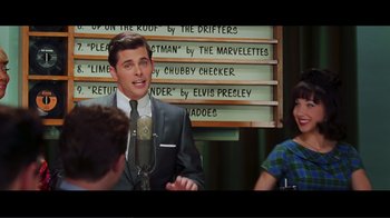Movie still from “Hairspray” (2007), directed by Adam Shankman – A man in a suit and tie talking into a microphone in front of two other people; Medium shot, Over the shoulder angle